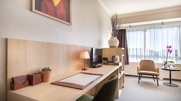 Quarto de hotel com uma escrivaninha de madeira, televisão, máquina de café, cadeira, pequena mesa com uma planta e janela com cortinas.