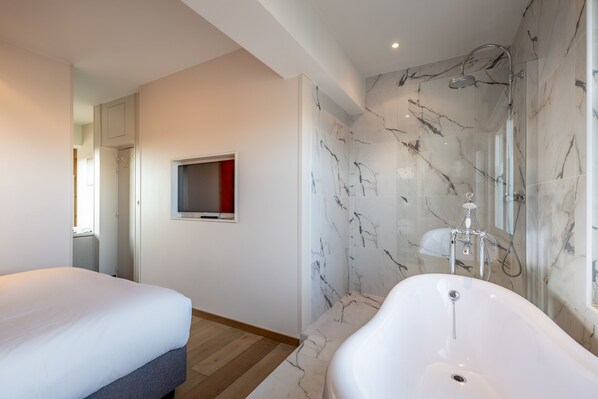 Una habitación de hotel moderna con una bañera grande, un televisor montado en la pared y una zona de dormitorio separada.