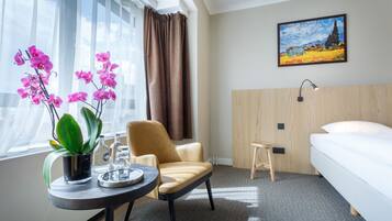 A hotel room with a bed, a chair, a small table with a plant, a painting on the wall, and a window with curtains.