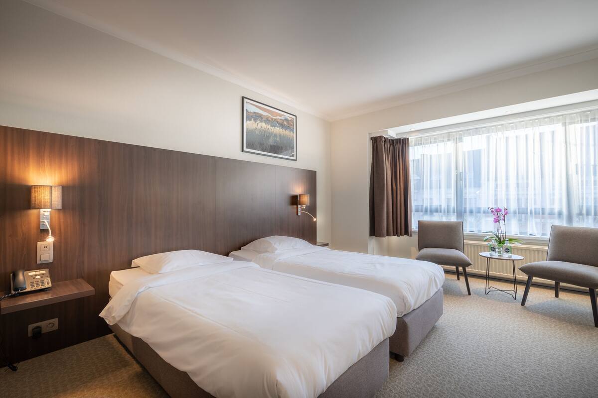 Ein Hotelzimmer mit zwei Betten, einer Sitzecke mit Tisch und Stühlen, einem Telefon und einem Fenster mit Vorhängen.