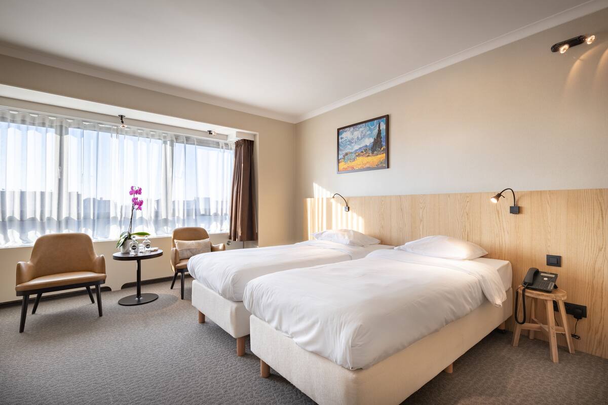 Ein Hotelzimmer mit zwei Betten, einem kleinen Tisch mit Pflanze, einem Sessel, einem Schreibtisch mit Telefon und einem Fenster mit Vorhängen.