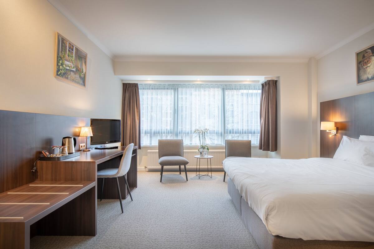 Ein Hotelzimmer mit Bett, Schreibtisch, Stühlen, einem kleinen Tisch mit Lampe und einem Fernseher.