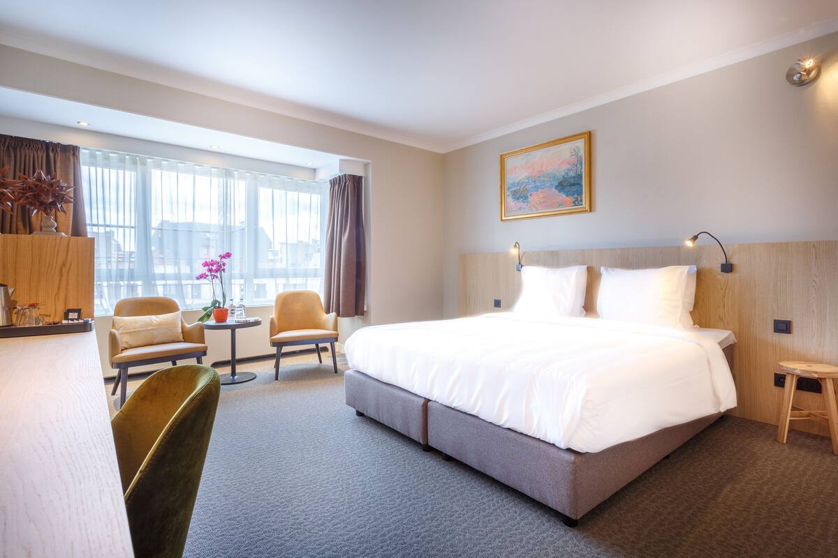 Ein Hotelzimmer mit einem großen Bett, einem Schreibtisch, zwei Stühlen, einem kleinen Tisch und einem Fenster mit Vorhängen.