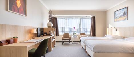 Habitación de hotel con cama, escritorio, silla y una pequeña mesa con lámpara. Se ve una ventana con cortinas y vistas al exterior.