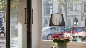 Una habitación con un ventanal grande, un florero con flores y una lámpara sobre una mesita.