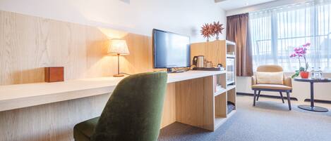 A hotel room with a desk, a green chair, a white chair, a television, a lamp, a bookshelf, a coffee maker, and a window with curtains.