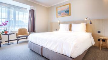 A hotel room with a large bed, a small table with a chair, a potted plant, and a painting on the wall.