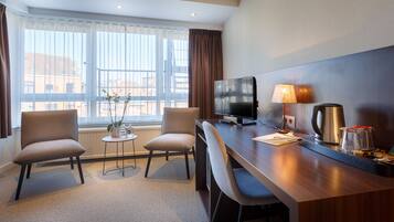 Une chambre d’hôtel dotée d’une grande fenêtre, d’un bureau avec un téléviseur, de deux chaises, d’une petite table avec une plante et d’une machine à café.