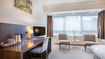 Ein Hotelzimmer mit Schreibtisch, zwei Stühlen, einem Bett, einem Fernseher, einer Kaffeemaschine und einem Fenster mit Vorhängen.
