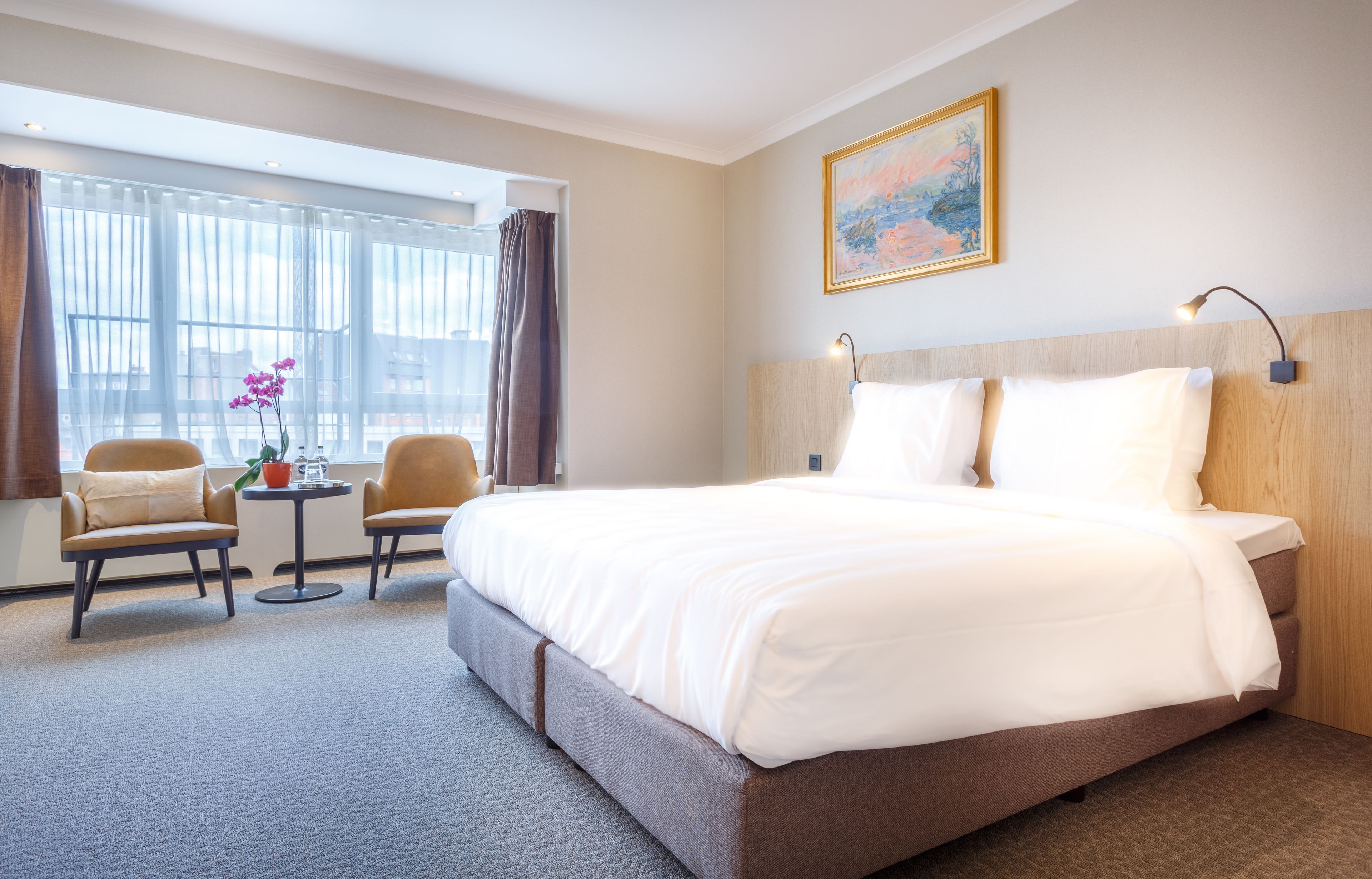 A hotel room with a large bed, two armchairs, a small table with a vase of flowers, and a large window with curtains.