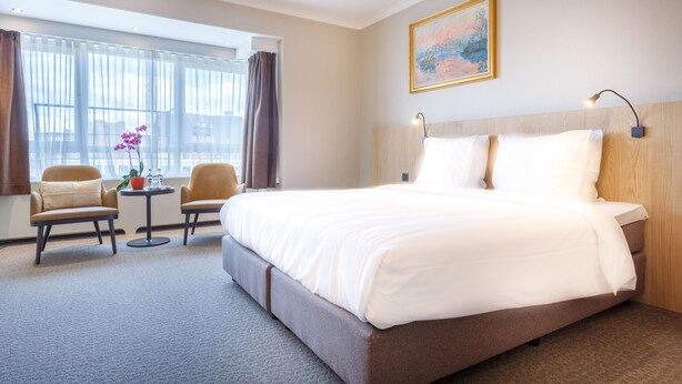 A hotel room with a large bed, two armchairs, a small table with a vase of flowers, and a large window with curtains. - Carlton Hotel (Ghent)