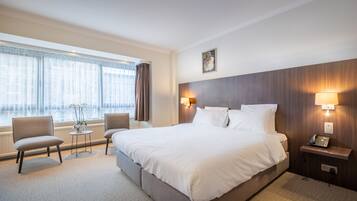Habitación de hotel con una cama grande, dos sillones, una mesita con un florero, y una mesita de noche con una lámpara y teléfono.