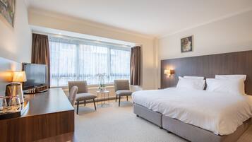 Habitación de hotel con una cama grande, un escritorio con televisor, dos sillas, una mesita con lámpara y una ventana con cortinas.