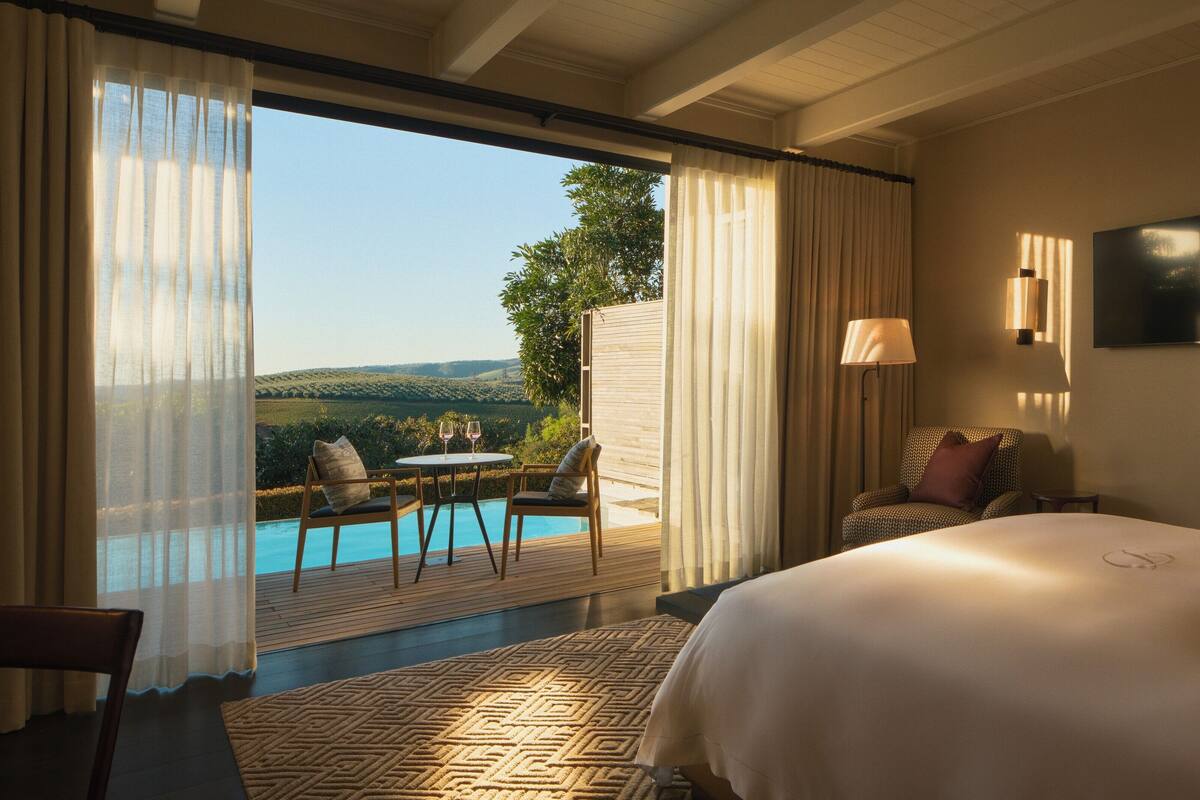 Quarto de hotel com cama, televisão, vista para a piscina e área de refeições com cadeiras e mesa.