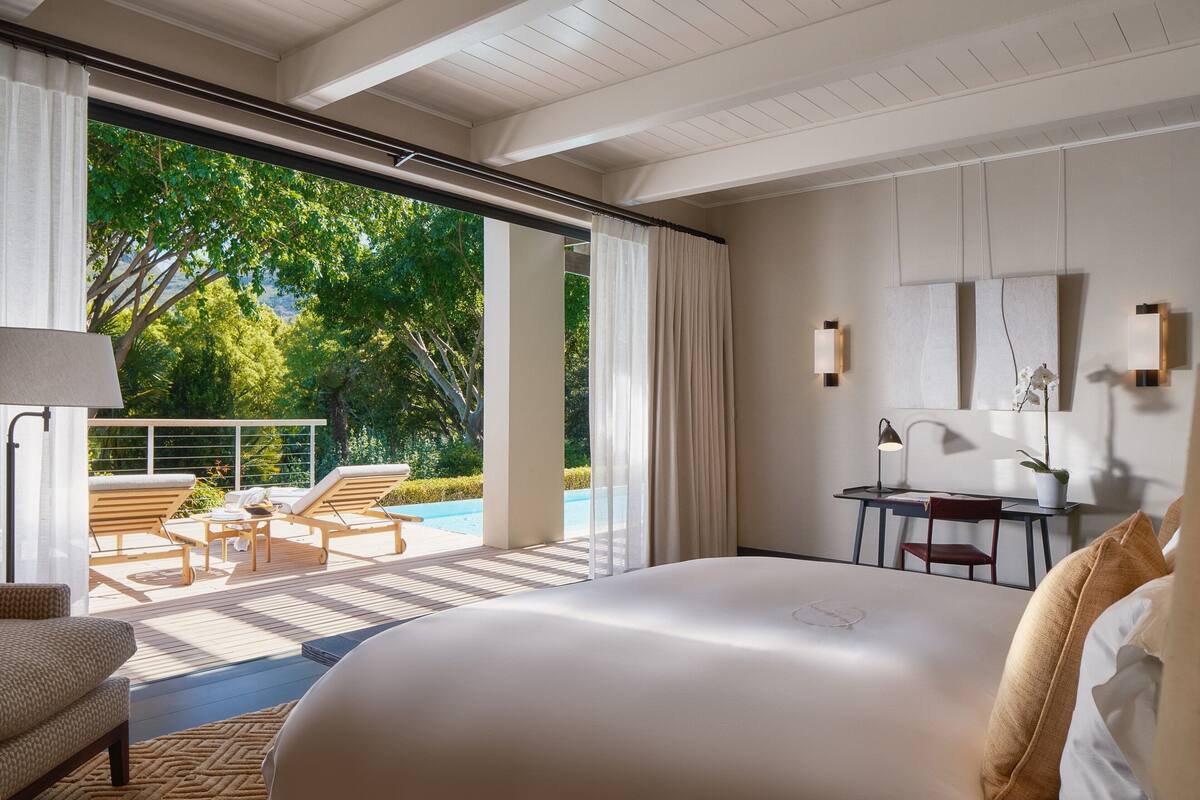 Quarto de hotel moderno com uma cama grande, uma escrivaninha e vista para uma piscina e vegetação lá fora.