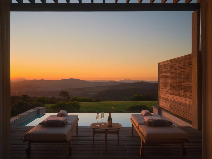 A wooden deck with two lounge chairs and a table, overlooking a scenic landscape at sunset.