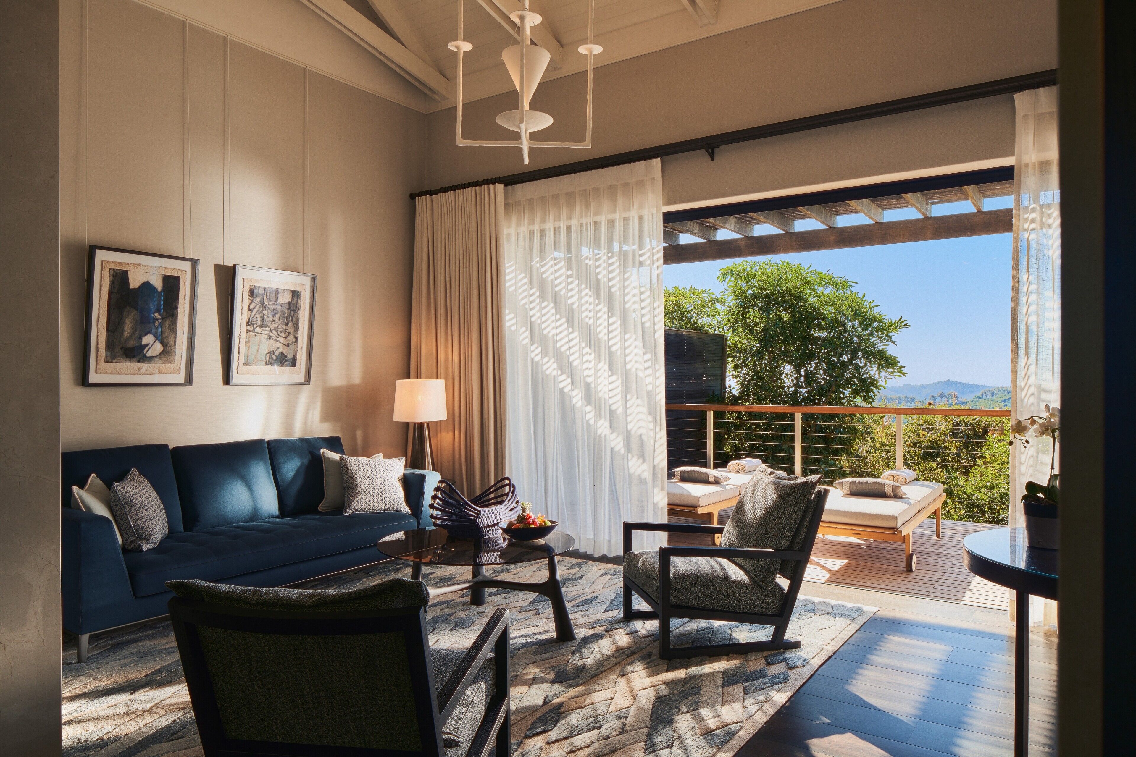 A modern living room with a dark blue sofa, a round coffee table, and a comfortable chair. There are two framed artworks on the wall, a large window with sheer curtains, and a balcony with outdoor seating.