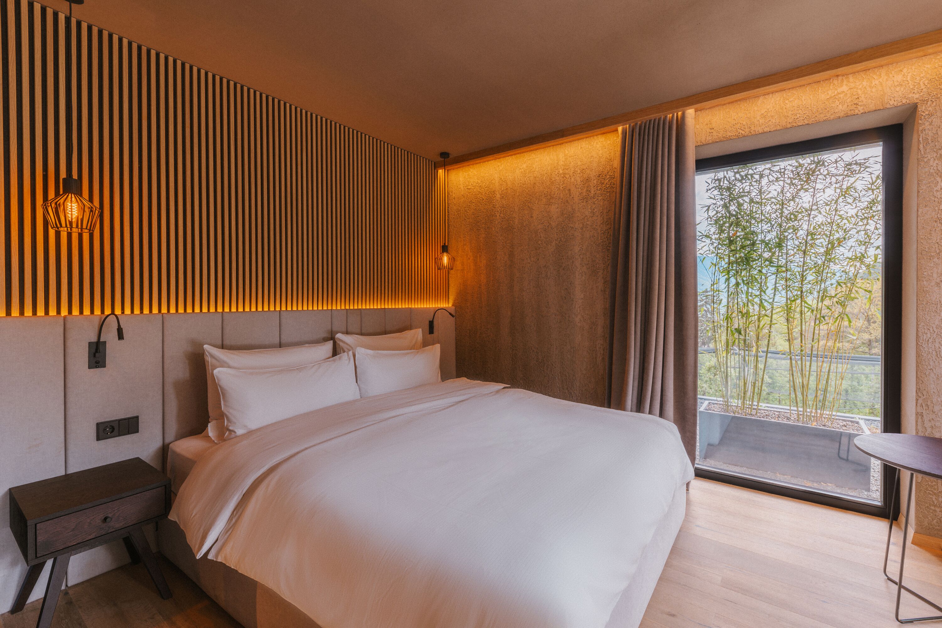 Habitación de hotel moderna con una cama grande, una mesita de noche, una pequeña mesa junto a la ventana y vistas a zonas verdes.