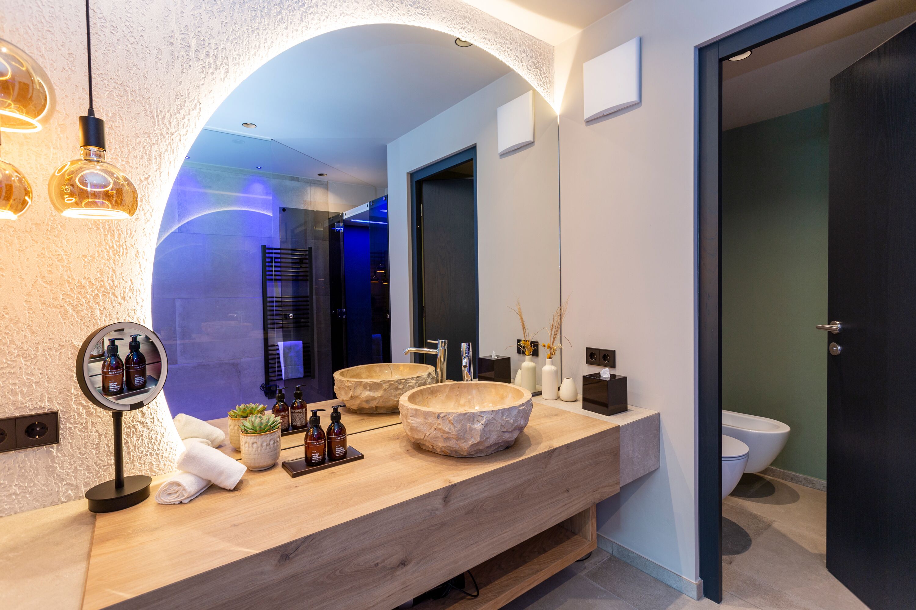 A modern bathroom with a wooden vanity, two large stone sinks, a round mirror, and a dark door.