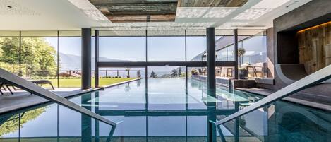 A modern indoor pool with glass railings, large windows offering a view of the outdoors, and a wooden ceiling.