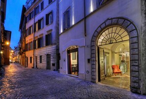 Une rue pavée mène à un bâtiment doté d’une grande entrée en arc de cercle, et on peut apercevoir à l’intérieur une chaise et une table.