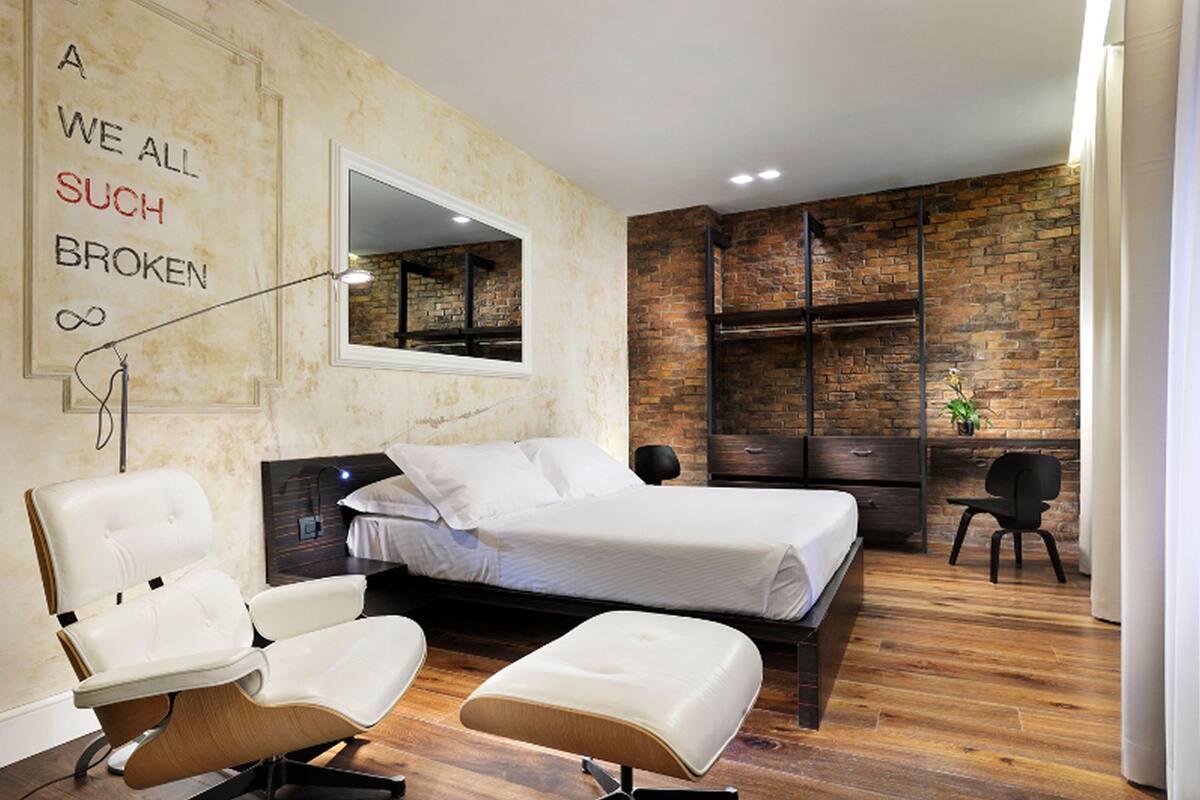 Un dormitorio moderno con pared de ladrillo, una cama grande, una silla blanca, una mesita y un espejo.