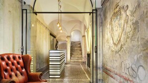 A hallway with a leather armchair, a stack of books, and a staircase in the background. - Relais Orso (Rome)