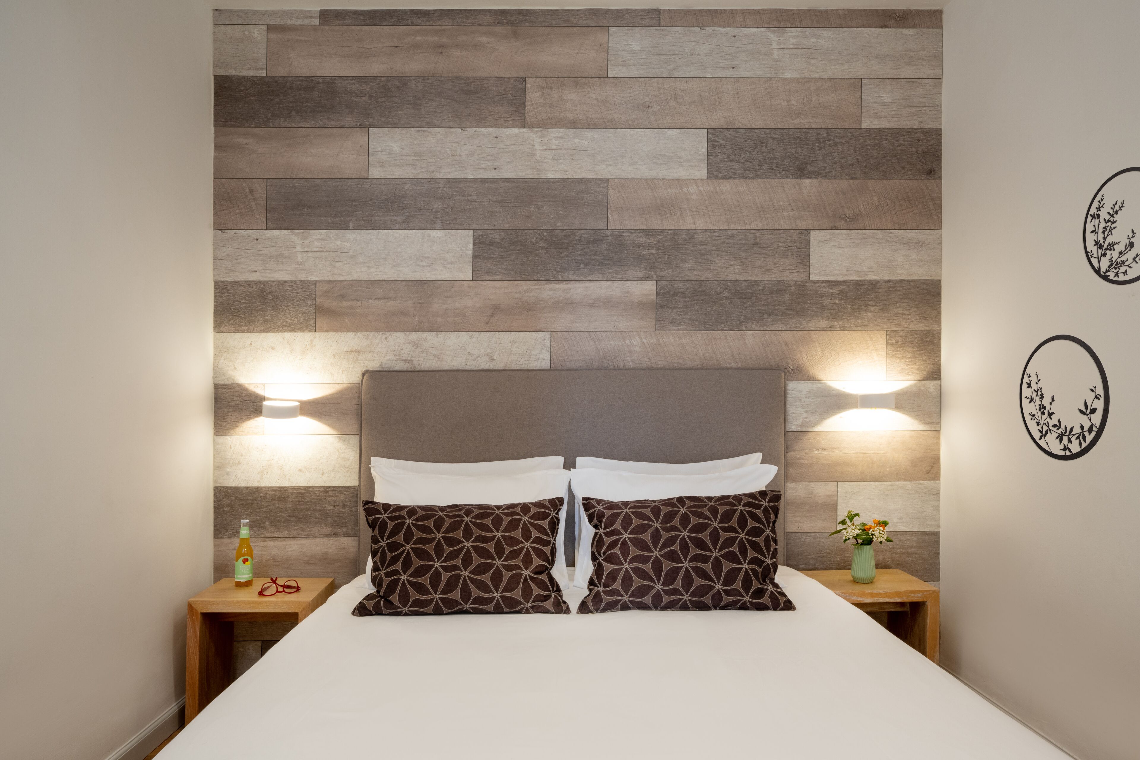 A neatly made bed with patterned pillows, a wooden headboard, and bedside tables with lamps and a bottle.