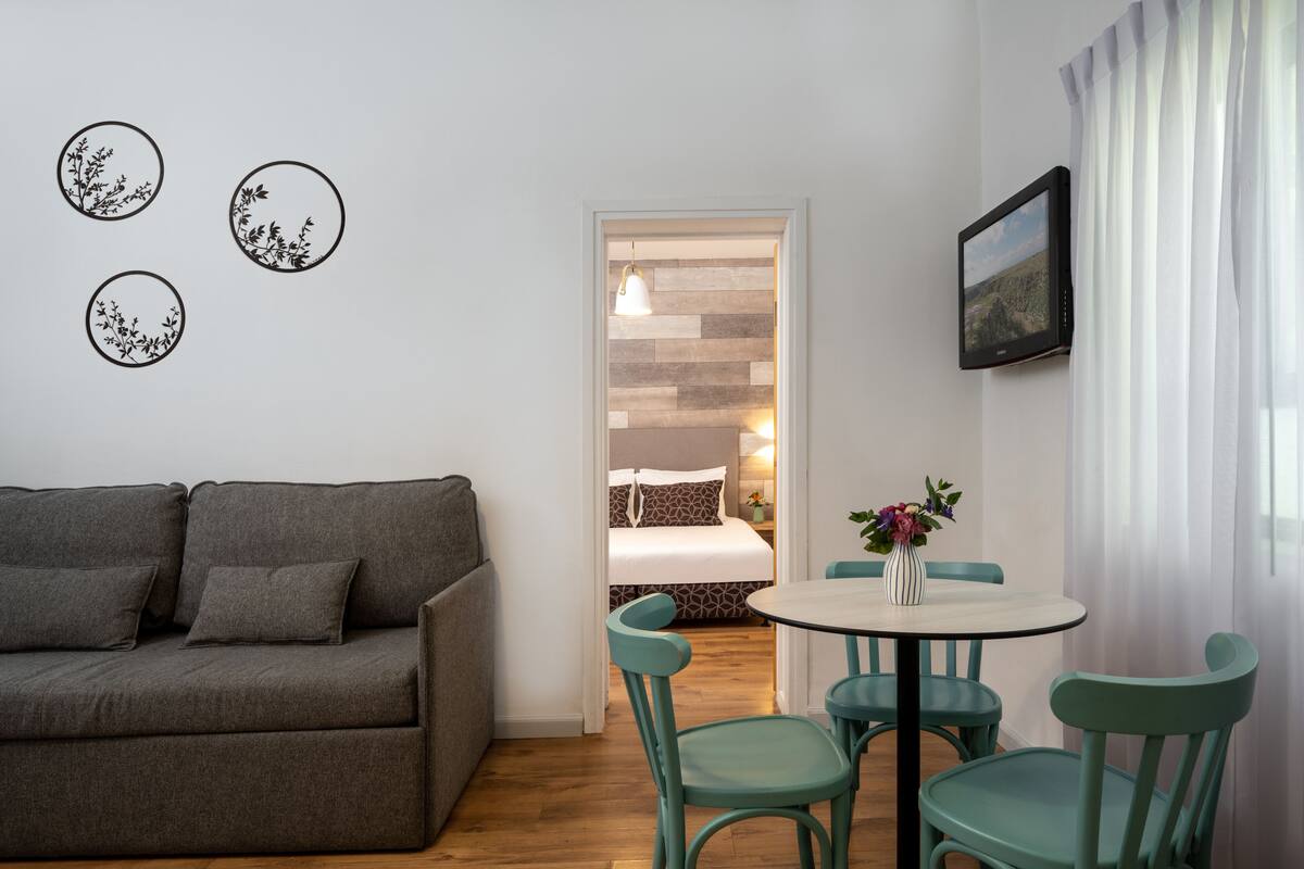 Une pièce chaleureuse avec un canapé gris, une table ronde entourée de chaises, un lit doté d’une tête de lit en bois et un tableau encadré accroché au mur.