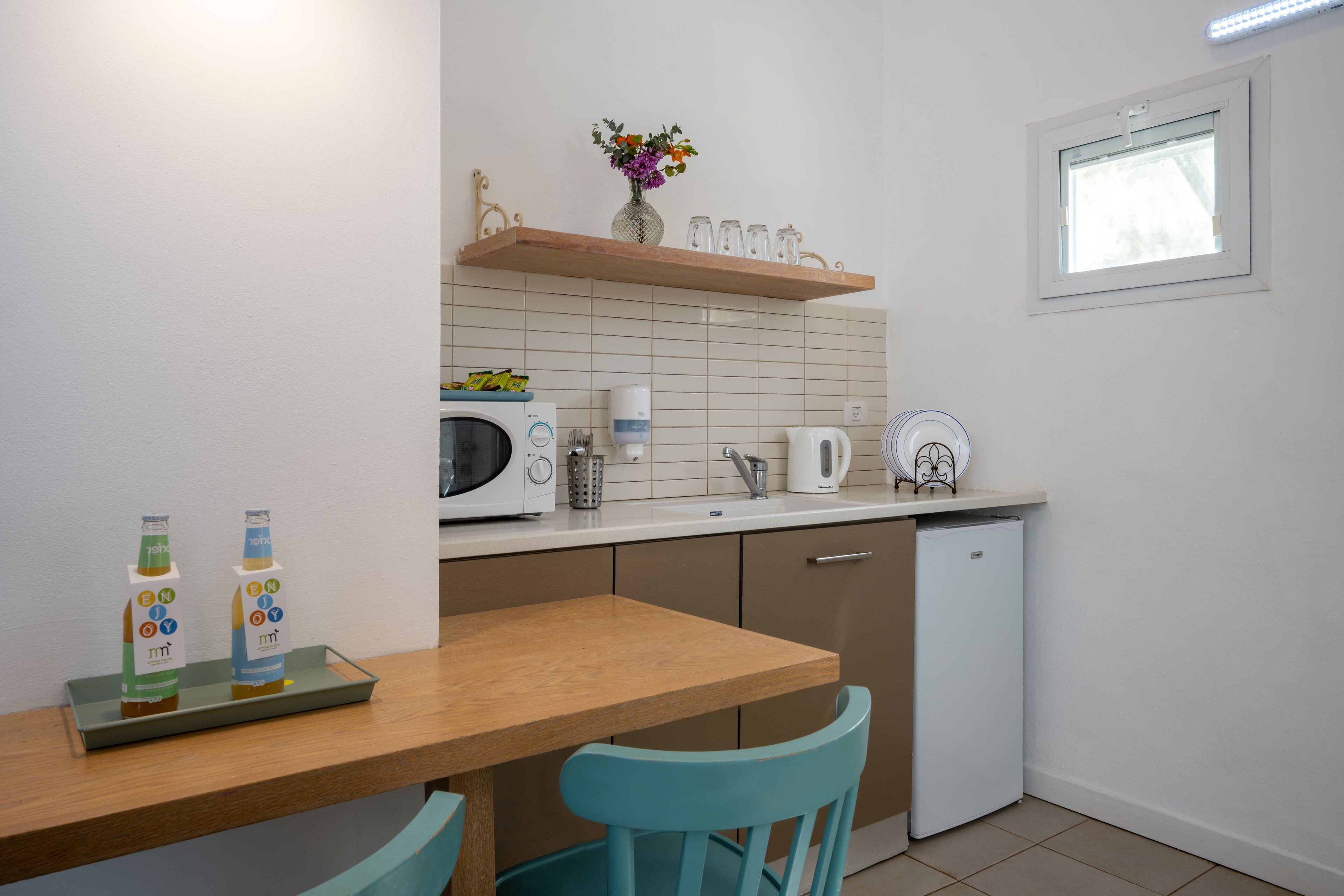 A compact kitchen with a microwave, toaster, and small refrigerator. There is a wooden table with two chairs and a tray with juice bottles. A vase with flowers is on a shelf above the counter.
