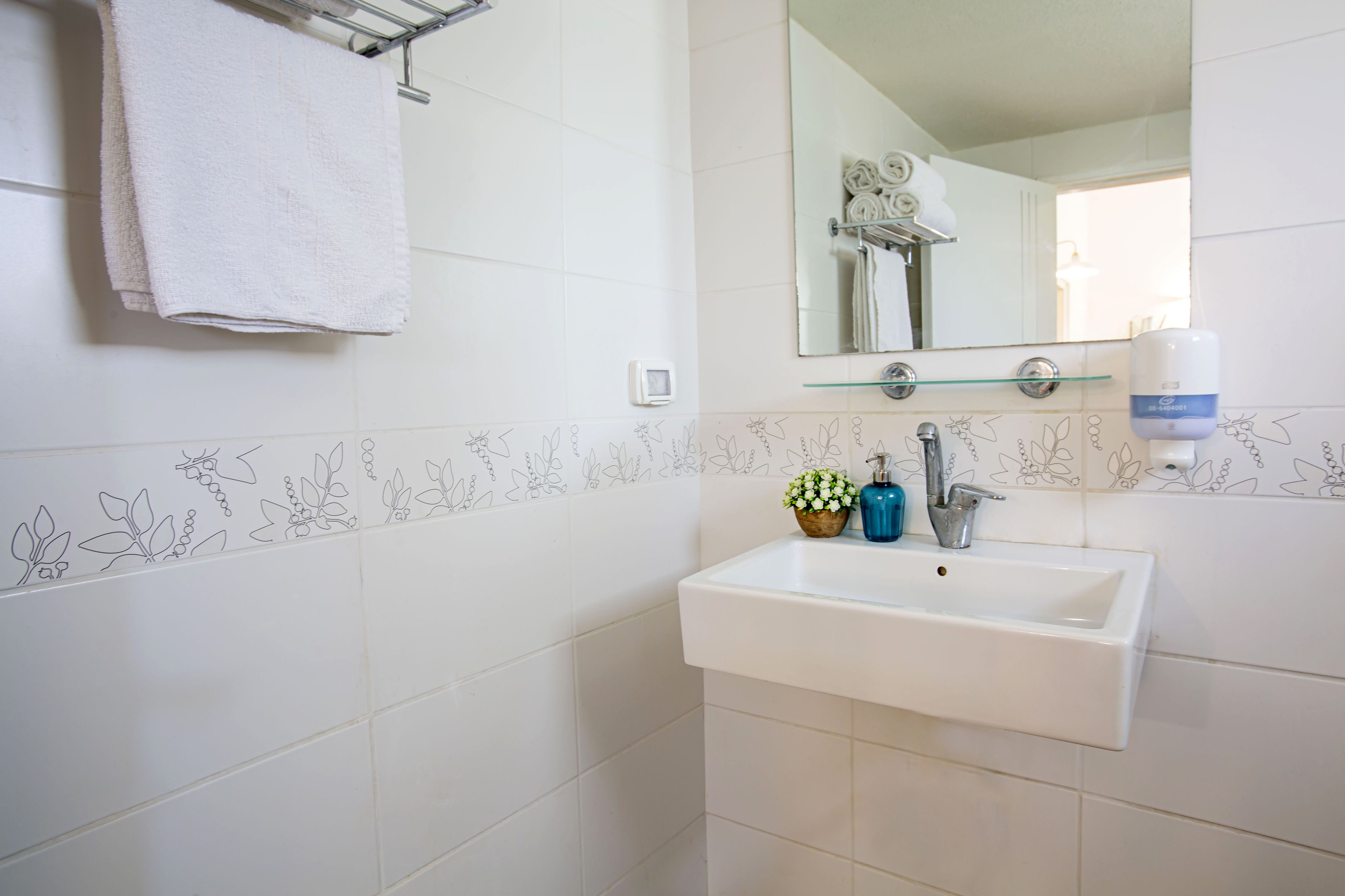Casa de banho com azulejos brancos, uma pia branca, espelho, suporte para toalhas, dispensador de sabonete e uma pequena planta.