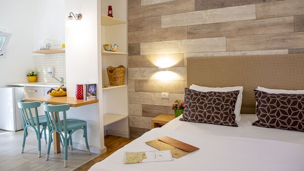 Una habitación de hotel moderna con cabecera de madera, cama con almohadones estampados, escritorio con sillas y una zona de cocina con refrigerador y estanterías.
