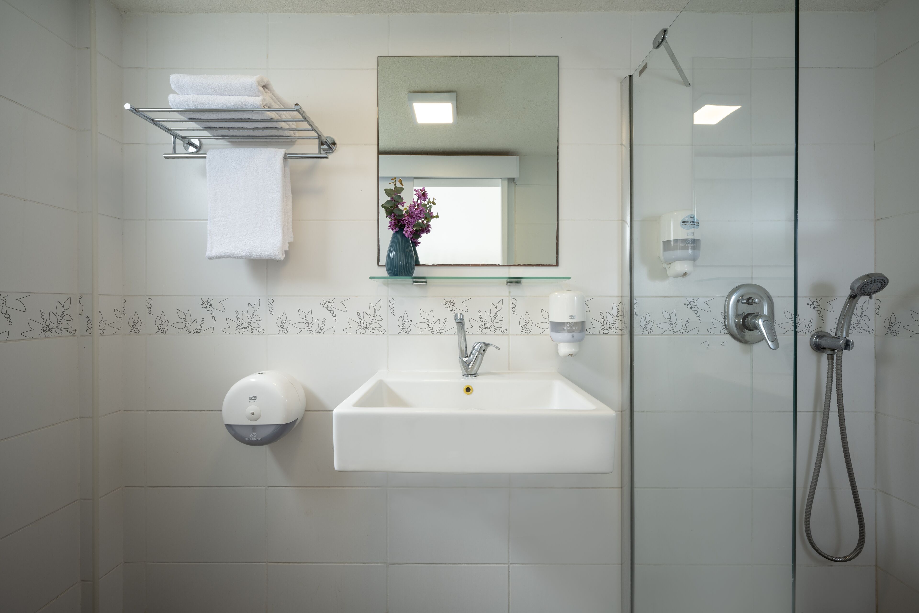 A bathroom with a white sink, a glass shower enclosure, a towel rack with a white towel, a mirror, and a vase with purple flowers.