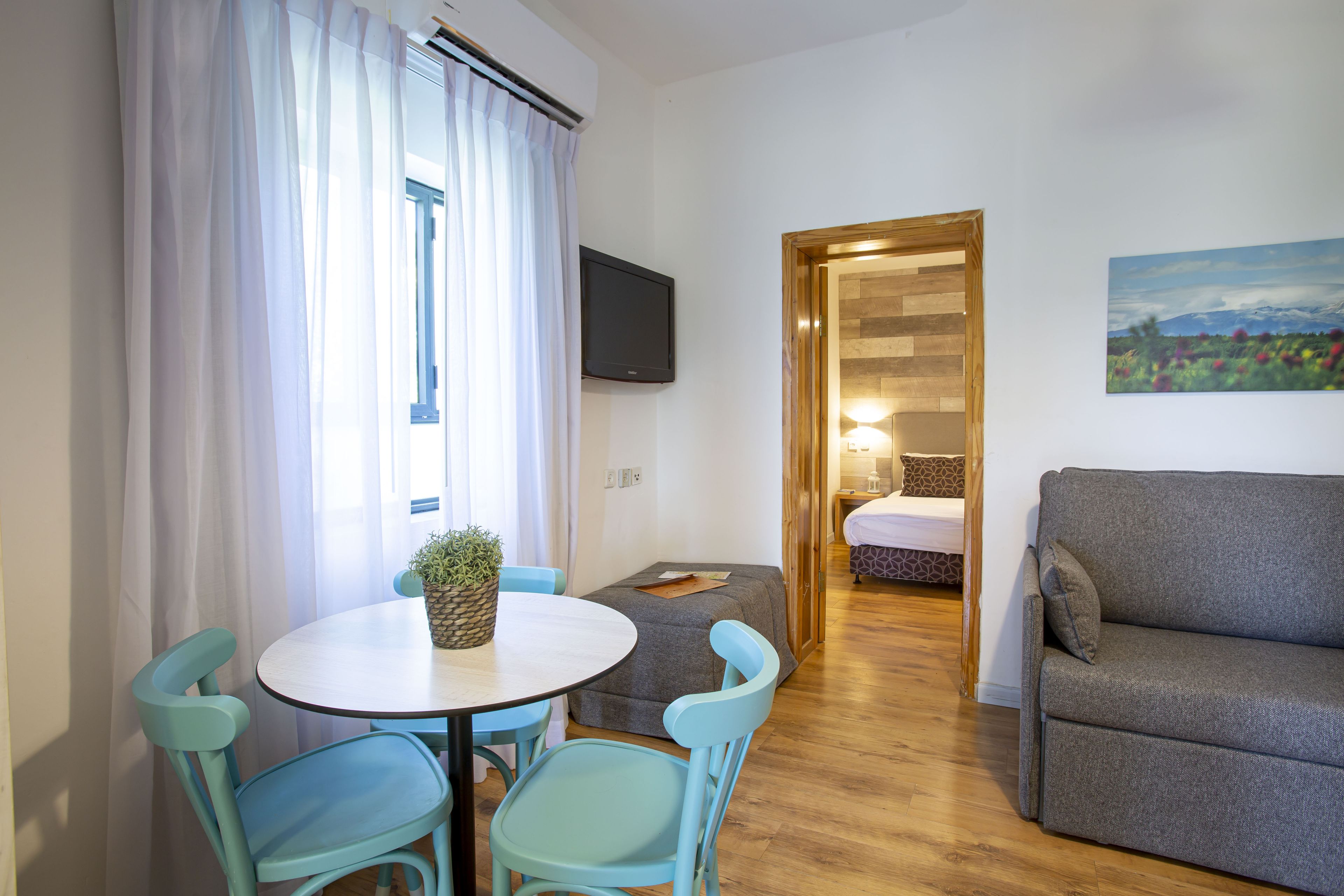 A room with a round table, two chairs, a sofa, a TV mounted on the wall, and a painting.