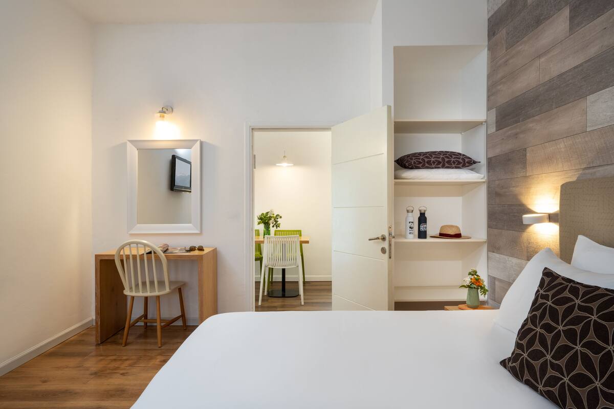 Une chambre d’hôtel moderne, équipée d’un lit, d’un bureau et d’une chaise. On peut apercevoir la salle de bain à travers une porte ouverte.