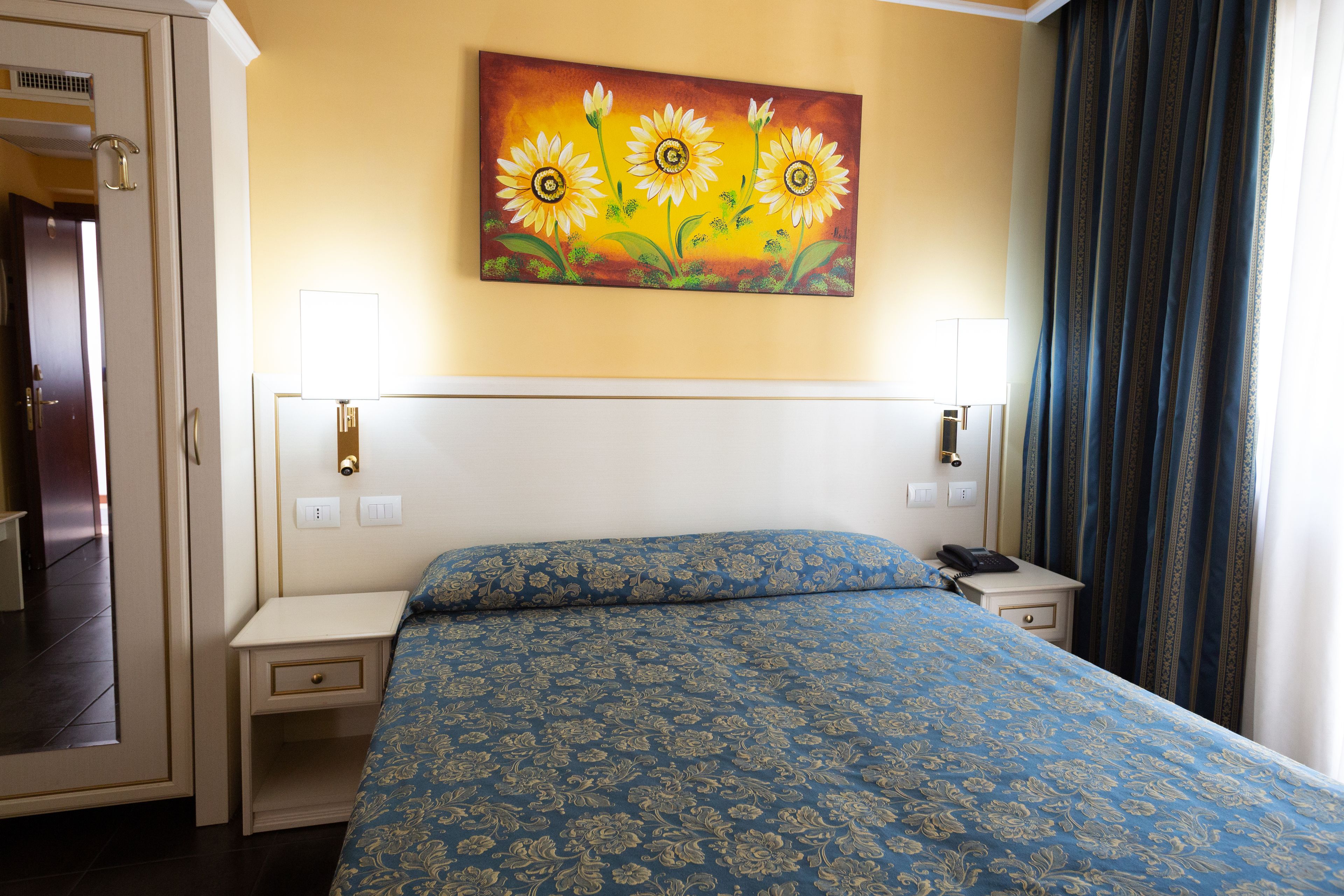 Ein Hotelzimmer mit einem großen Bett, zwei Nachttischen, einem Schminktisch, einem Gemälde von Sonnenblumen und einem Fenster mit Vorhängen.