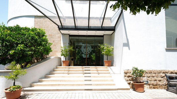 A building entrance with a staircase leading up to a glass door, flanked by potted plants. - Cervara Park Hotel (Rome)