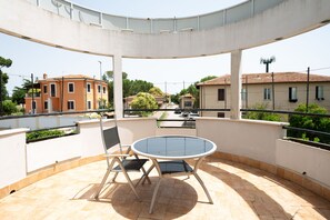 A covered outdoor patio with a round table and two chairs, overlooking a street with buildings and a body of water. - Cervara Park Hotel (Rome)