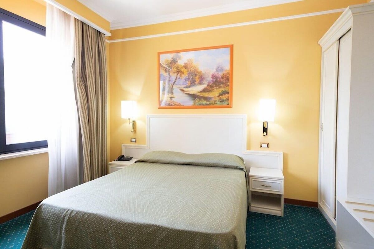 Ein Hotelzimmer mit einem Bett, zwei Nachttischen, einem Hängebild und einem Fenster mit Vorhängen.