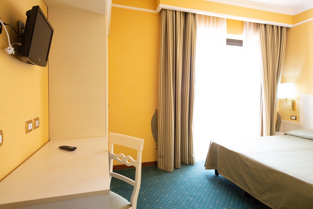 A hotel room with a bed, a desk, a chair, a television mounted on the wall, and curtains on the window.
