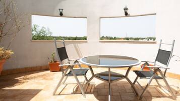 Une terrasse avec une table ronde et quatre chaises, deux fenêtres donnant sur un espace verdoyant et une plante en pot.