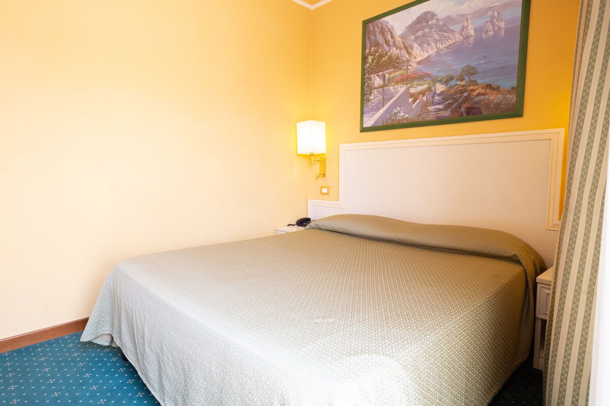 A hotel room with a bed, a nightstand, a lamp, and a painting on the wall.