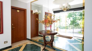 A lobby with a wooden door, a round table with a vase of flowers, and a large mirror reflecting the room. - Cervara Park Hotel (Rome)