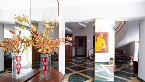 A lobby with a wooden reception desk, vases with dried leaves, a patterned floor, and a painting of a seated figure. - Cervara Park Hotel (Rome)