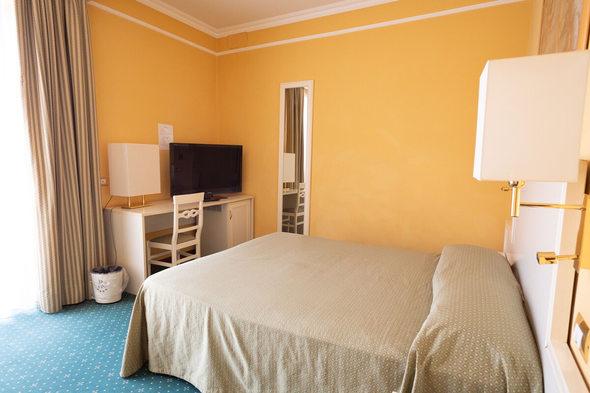 A hotel room with a bed, a desk with a computer and a TV, a mirror, and a window with curtains.