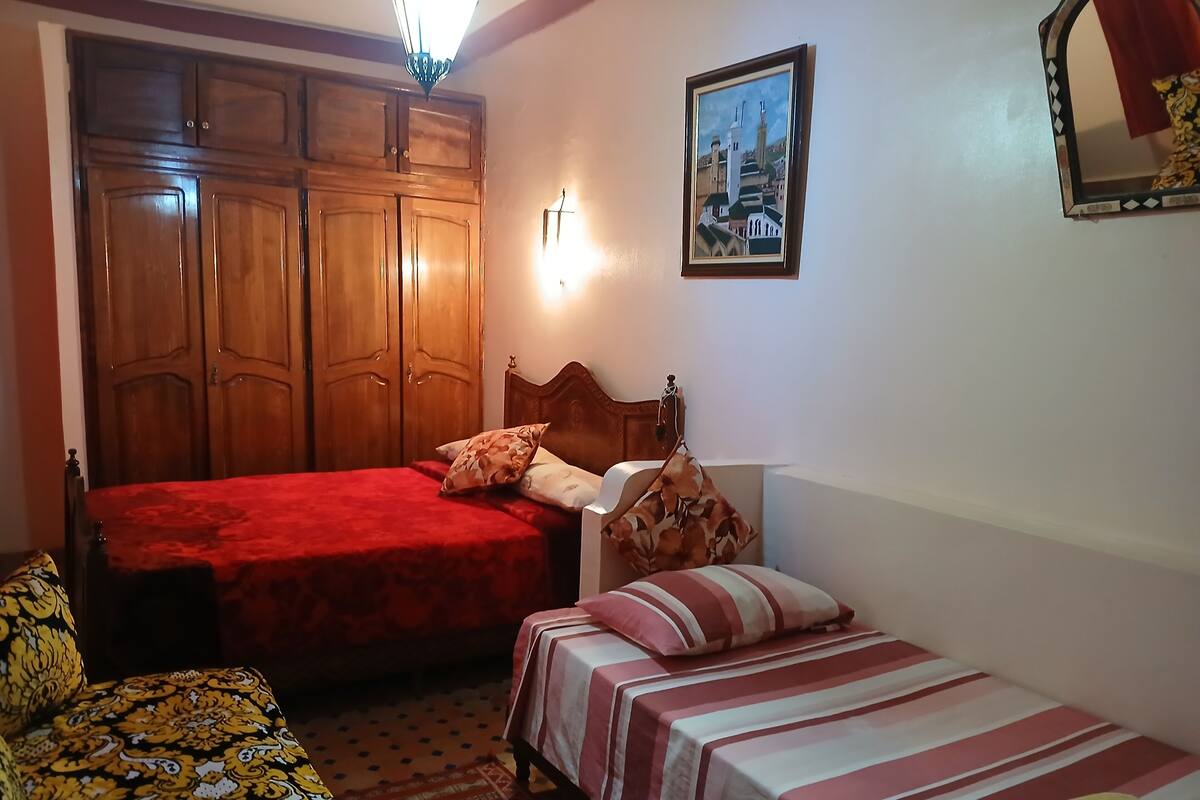 A bedroom with two beds, a wooden wardrobe, a wall-mounted lamp, and a framed picture.
