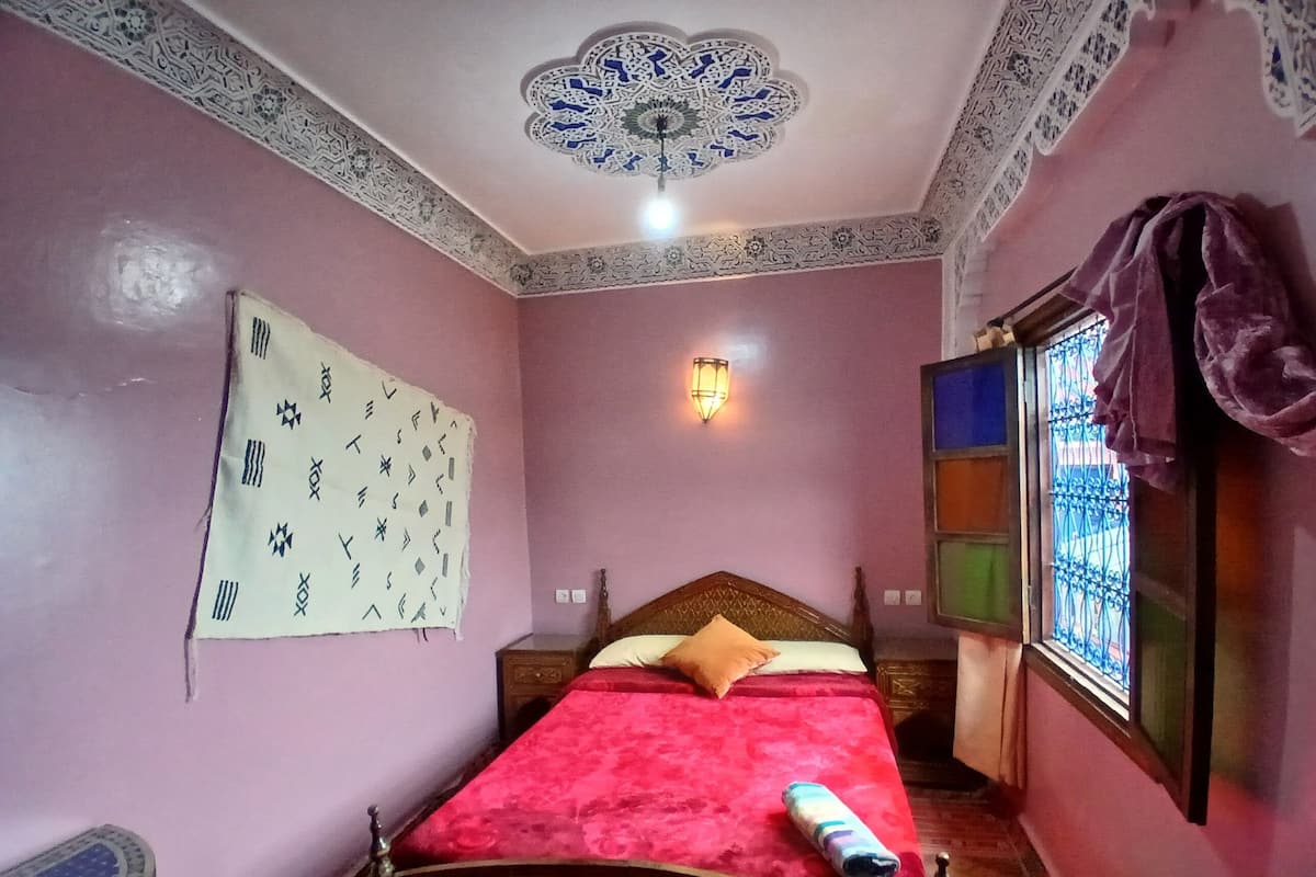 A bedroom with a bed, a wall-mounted light, a patterned wall hanging, and a window with bars.