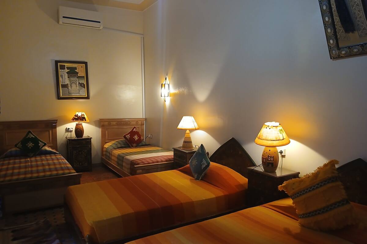 A hotel room with two single beds, a nightstand with a lamp, a wall-mounted air conditioner, and a framed picture on the wall.