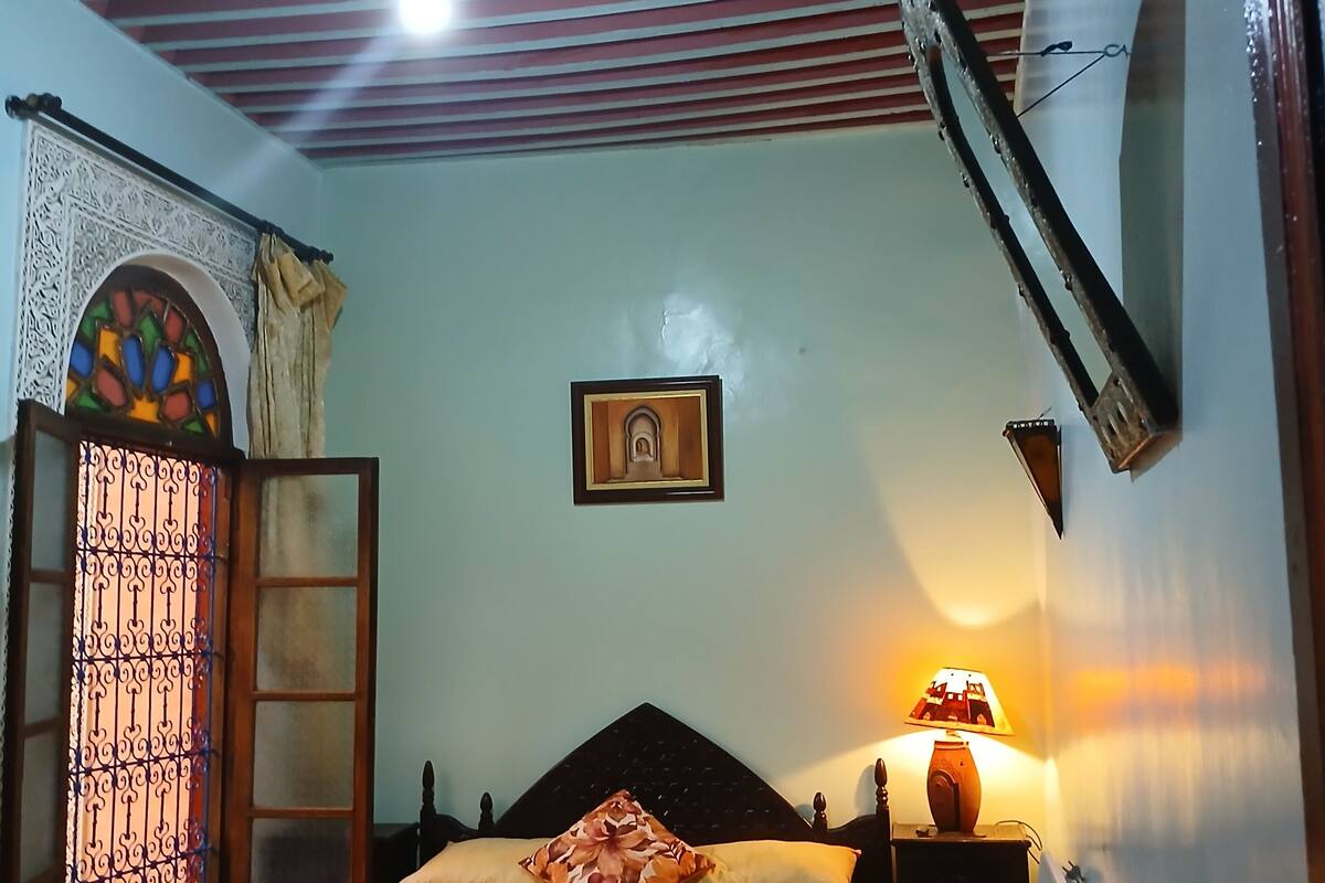 A room with a bed, a nightstand with a lamp, a decorative ceiling, and a window with a patterned screen.