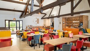 A modern dining area with wooden tables and colorful chairs, a kitchen in the background, and exposed beams on the ceiling.